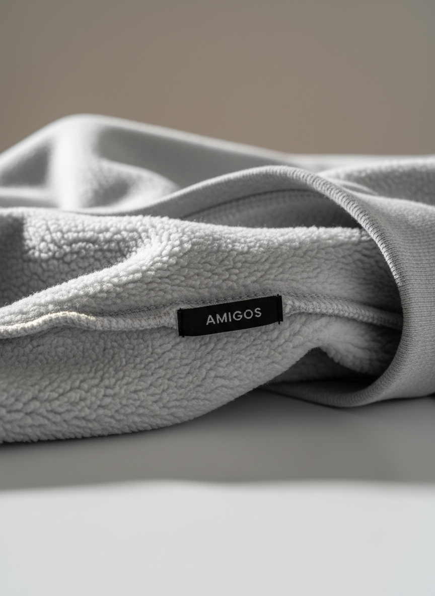 A refined Amigos brand detail shot focusing on a close-up of a sweatshirt’s interior fleece lining in warm light gray, its fibers appearing exceptionally soft and dense. The garment is draped over a smooth, matte white surface, with a neatly sewn Amigos label visible along the seam in understated black lettering. Soft studio lighting from the side creates delicate shadows within the fleece texture and subtle highlights along the clean stitching. The background falls gently out of focus, retaining only neutral tones, so all attention remains on the craftsmanship and comfort. Composed tightly with a macro-like perspective, the photographic style and minimal palette reinforce a premium, professional, comfort-first identity for the clothing brand.